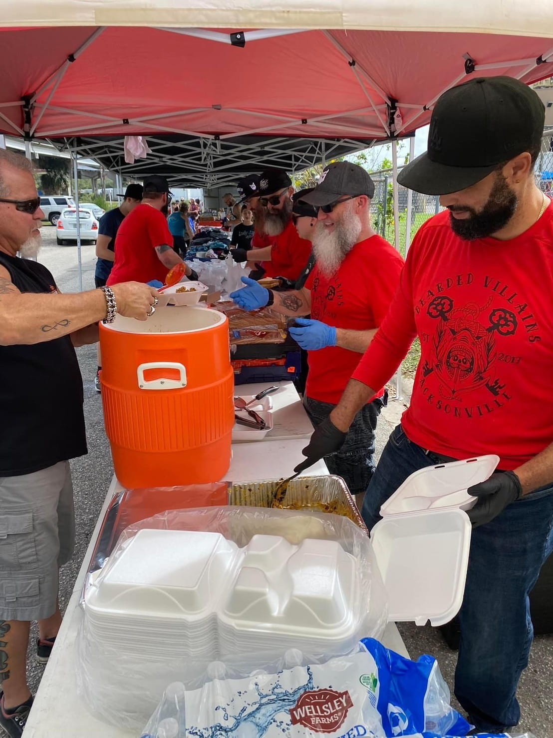 Bearded villains volunteers serving food to the homeless under a red awning. Villains are wearing the red bearded villains shirts worn for public events.